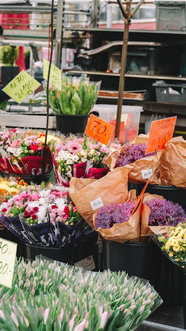 A vibrant display of fresh flower bouquets arranged outdoors at a flower market, showcasing a variety of colorful blossoms including pink, white, purple, and green flowers. The arrangements are wrappe