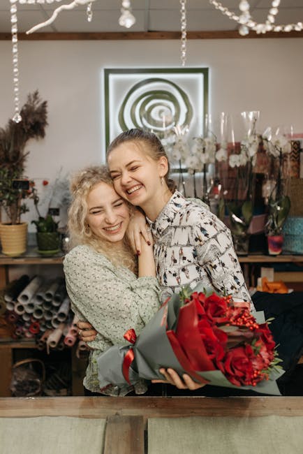 Two women stand close together inside a florist shop, smiling joyfully. One woman, with curly blonde hair, wears a light green patterned blouse, while the other, with straight brown hair, wears a whit