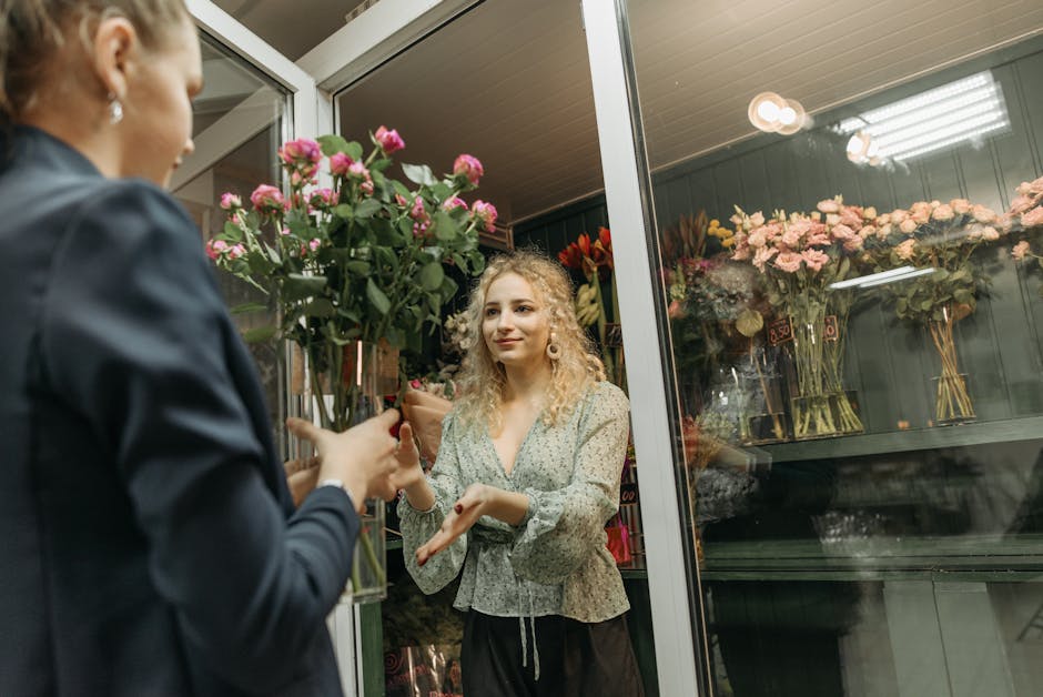A young woman with curly blonde hair, wearing a light-colored floral blouse, is standing inside a florist shop near a large glass display case filled with various floral arrangements and bouquets, som