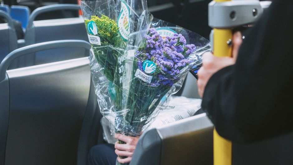 A person holding a bouquet of fresh purple and green flowers wrapped in clear plastic, with visible labels indicating the floral arrangement is from a professional florist. The bouquet includes purple