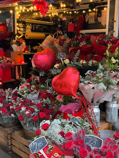 A vibrant display of fresh floral arrangements at a flower shop, featuring predominantly red roses, white blossoms, and greenery, accented with large red heart-shaped balloons. The flowers are arrange