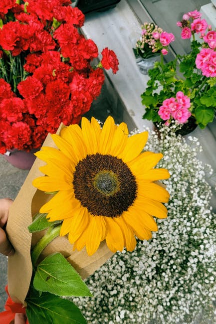 A vibrant bouquet featuring a large sunflower with bright yellow petals and a dark brown center, wrapped in brown craft paper. The bouquet is held by a person whose hand is partially visible. In the b