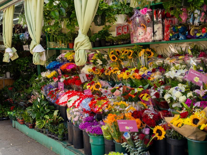 A vibrant floral display outside a flower shop features a variety of fresh bouquets arranged on tiered tables. The arrangement includes sunflowers with bright yellow petals and dark centers, mixed wit