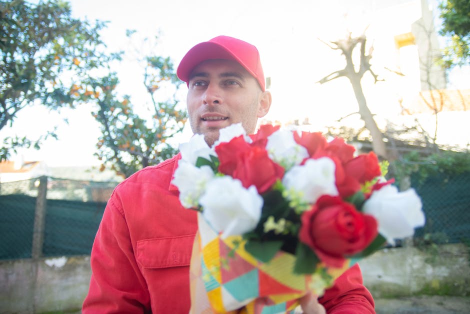 A man wearing a red cap and matching red jacket is holding a vibrant bouquet of fresh flowers, including red and white roses, wrapped in colorful patterned paper. He is outdoors, with blurred green tr