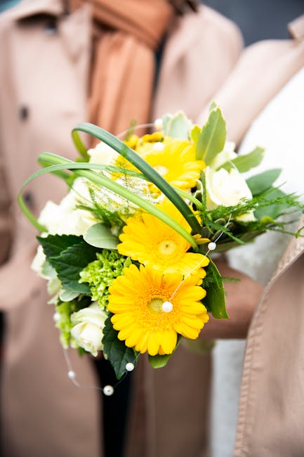 A floral arrangement featuring bright yellow Gerbera daisies, complemented by white roses and small green hypericum berries, all arranged with lush green foliage including large leaves and delicate fe