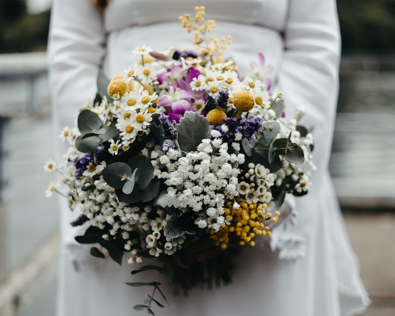 A person dressed in a white outfit is holding a large bouquet of fresh flowers, displayed directly in front of them. The arrangement includes white daisies, small yellow button poms, delicate purple o