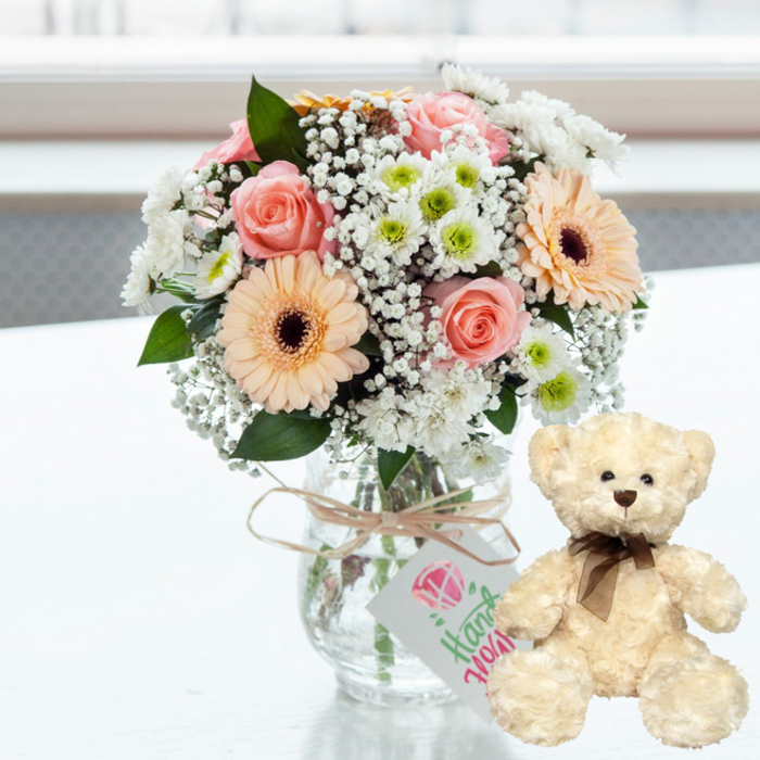 A soothing, celebratory scene is captured on a clean white surface, where a pastel bouquet in a glass jar shares space with a small cream teddy bear, both bathed in airy, natural light reminiscent of a Kentish Town terrace on a clear day. The bouquet is arranged in a round, compact style, yet every stem feels intentionally placed. Soft pink roses, just opening, are woven among peach gerbera daisies whose dark brown centres form a warm focal point. Around them, small white chrysanthemums with hints of lime-green at their cores and delicate sprays of white baby's breath create a halo of lighter texture and colour. The clear glass jar-style vase reveals the bundled green stems, grounding the softness of the blooms in something simple and real. At the neck of the vase, a length of natural twine is wrapped and tied, holding a small, rectangular white tag adorned with a pink heart symbol and flowing green script, hinting that this arrangement is meant for someone special. To the right, a plush teddy bear in a creamy shade sits upright on the same reflective surface. Its fur appears thick and downy, the kind you instinctively want to touch, with dark bead eyes, a tiny brown nose, and a slim brown satin ribbon tied carefully in a bow at its neck. The background dissolves into a soft blur of pale tones, suggesting an out-of-focus window or wall, keeping the viewer's attention on the intimacy of the gift. It feels like a thoughtful anniversary arrangement a local Kentish Town florist might send to a home near Parliament Hill, blending romance, tenderness, and a gentle sense of home.