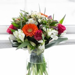 A radiant floral arrangement, brimming with local charm, commands attention in a tall glass vase, reflecting the spirited character of Kentish Town's neighbourhoods. Creamy peach gerberas gently frame the bouquet, their petals forming a soft halo that encircles bold orange centers-each like a beacon at the heart of a celebration. The deep magenta roses build rich contrast and a touch of luxury, their unfurling petals reminiscent of blooms just picked from a well-kept garden near Gospel Oak. Ivory alstroemeria and small white filler blossoms impart brightness and textural complexity, while blush-touched pink flowers introduce a playful lightness. Broad, paddle-shaped aspidistra leaves curve protectively around the base, anchoring the composition in lush tranquillity much like the leafy surrounds of Kentish Town's local parks. Smaller, sharp-tipped greens mingle with the flowers, filling the arrangement with vitality and movement. The clear vase beneath displays an organized profusion of stems, celebrating the natural structure and fresh-cut execution of a skilled local florist. Overhead, gentle light falls across the bouquet, emphasizing each colour's vibrancy and highlighting the convergence of artistry, freshness, and thoughtful arrangement. With a faint, rejuvenating scent in the air-a blend of roses, lilies, and crisp foliage-this ensemble is perfect for marking milestones, gathering friends on Kentish Town Road, or gifting to those embarking on a new journey. Every stem is placed with optimism and care, echoing the best of Kentish Town's floral tradition.